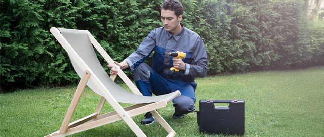 Man Repairing Wooden Deck Chair With Drill In Backyard Garden